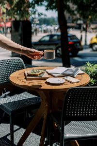 Woman placing coffee on bistro table with design magazines and laptop